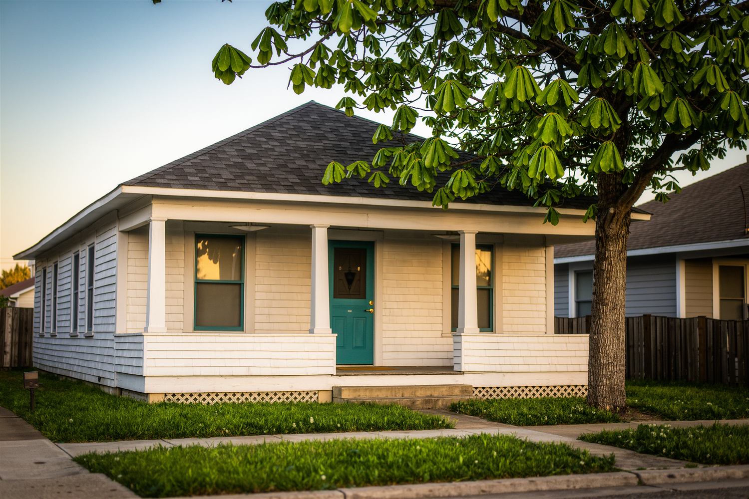 White bungalow with a covered front porch and a teal door beside a large shade tree