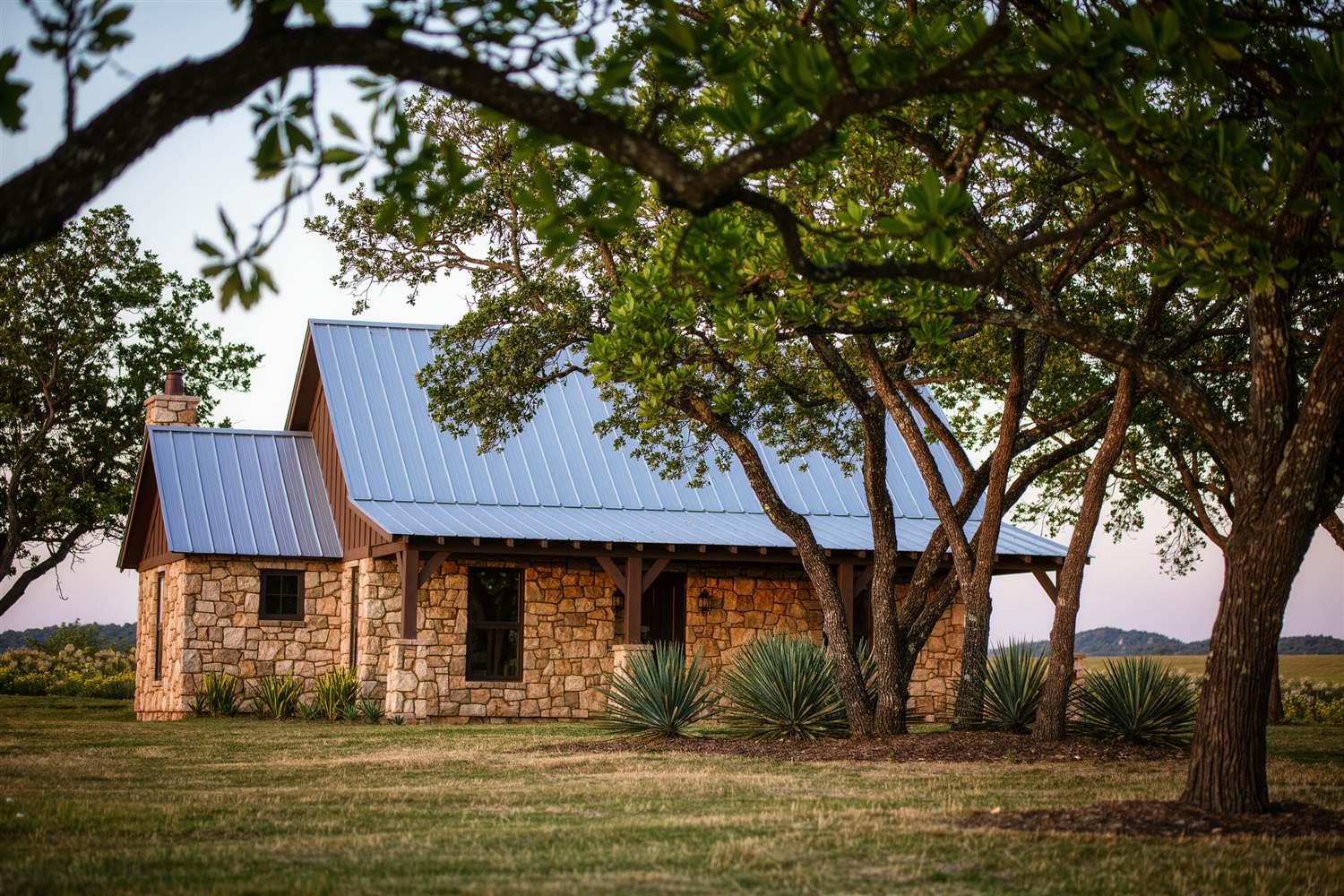 Stone ranch house with a metal roof among oaks and yucca plants
