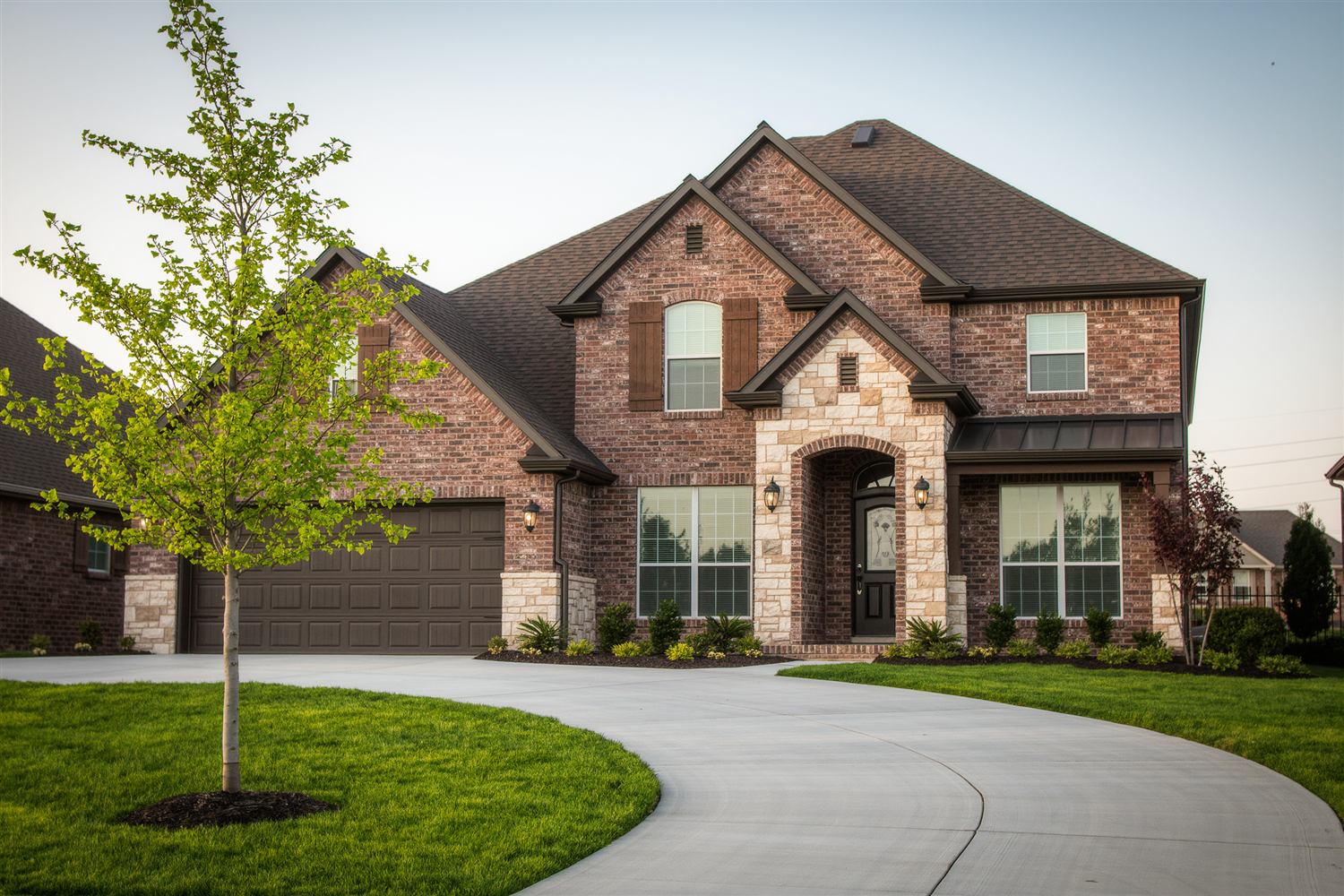 Two-story brick and stone house with a curved driveway and a young tree